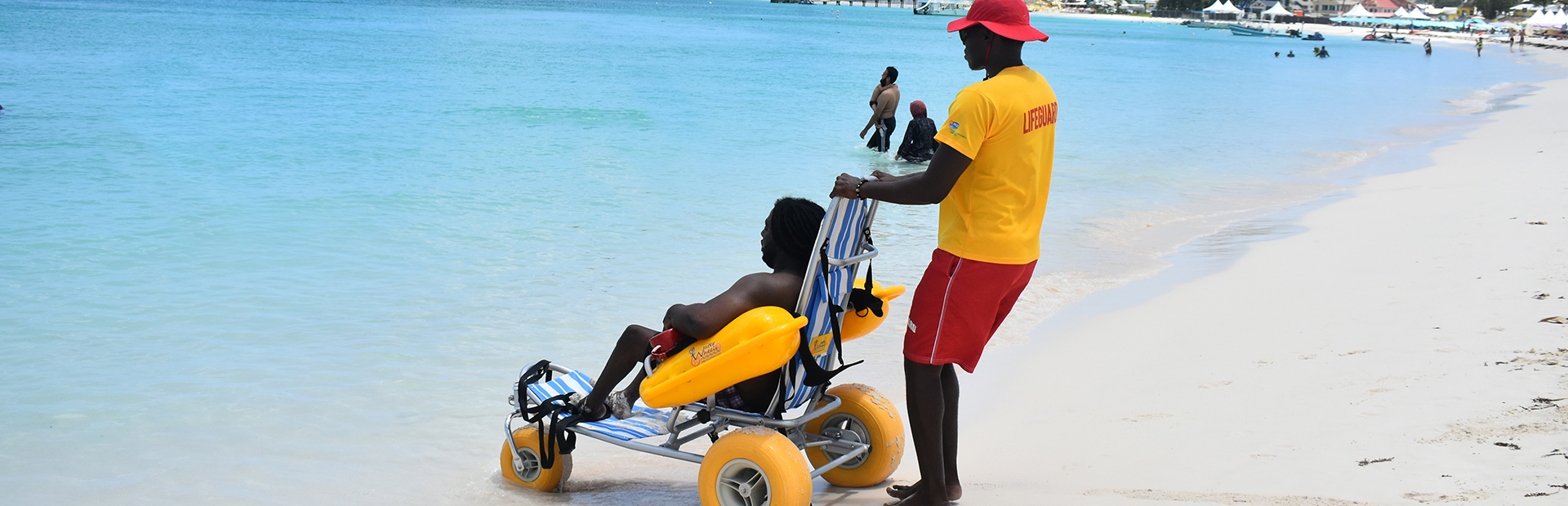 Young man in the Fully Accessible Beach wheelchair rented from the Barbados Council for the disabled being taken into the ocean by lifeguard on Brownes Beach in Barbados
